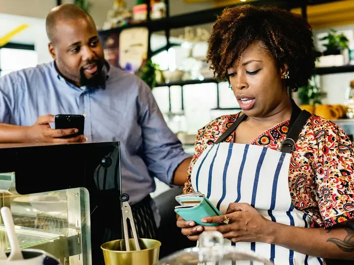 A woman in an apron using a phone near a cafe counter while a man watches, showing small business teamwork at Town Service Zone Now